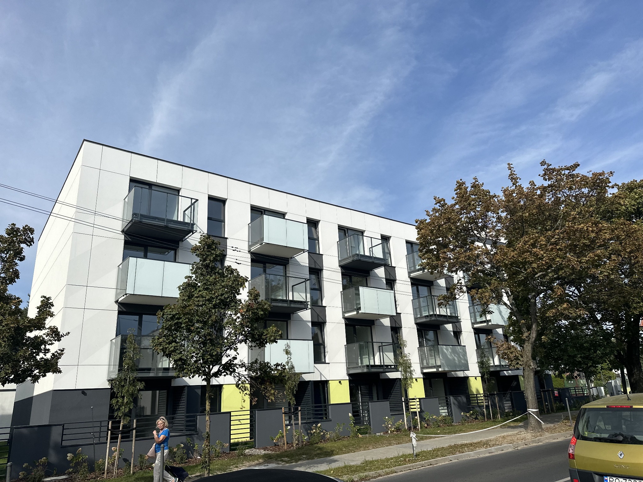 Nowoczesny budynek wielorodzinny z balkonami o szklanych balustradach, elewacja w kolorze białym i szarym, zieleń miejska przed budynkiem, kobieta z walizką na chodniku.