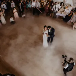 Para młoda w objęciach na parkiecie otoczonym gośćmi weselnymi i efektem mgły, filmowana z góry przez kamerzystę.