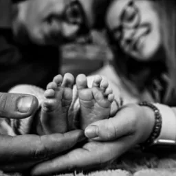 Czarno-białe zdjęcie dłoni rodzic&oacute;w trzymających stopy noworodka na tle rozmytych twarzy rodzic&oacute;w, mama ma na ręce bransoletkę i zegarek.