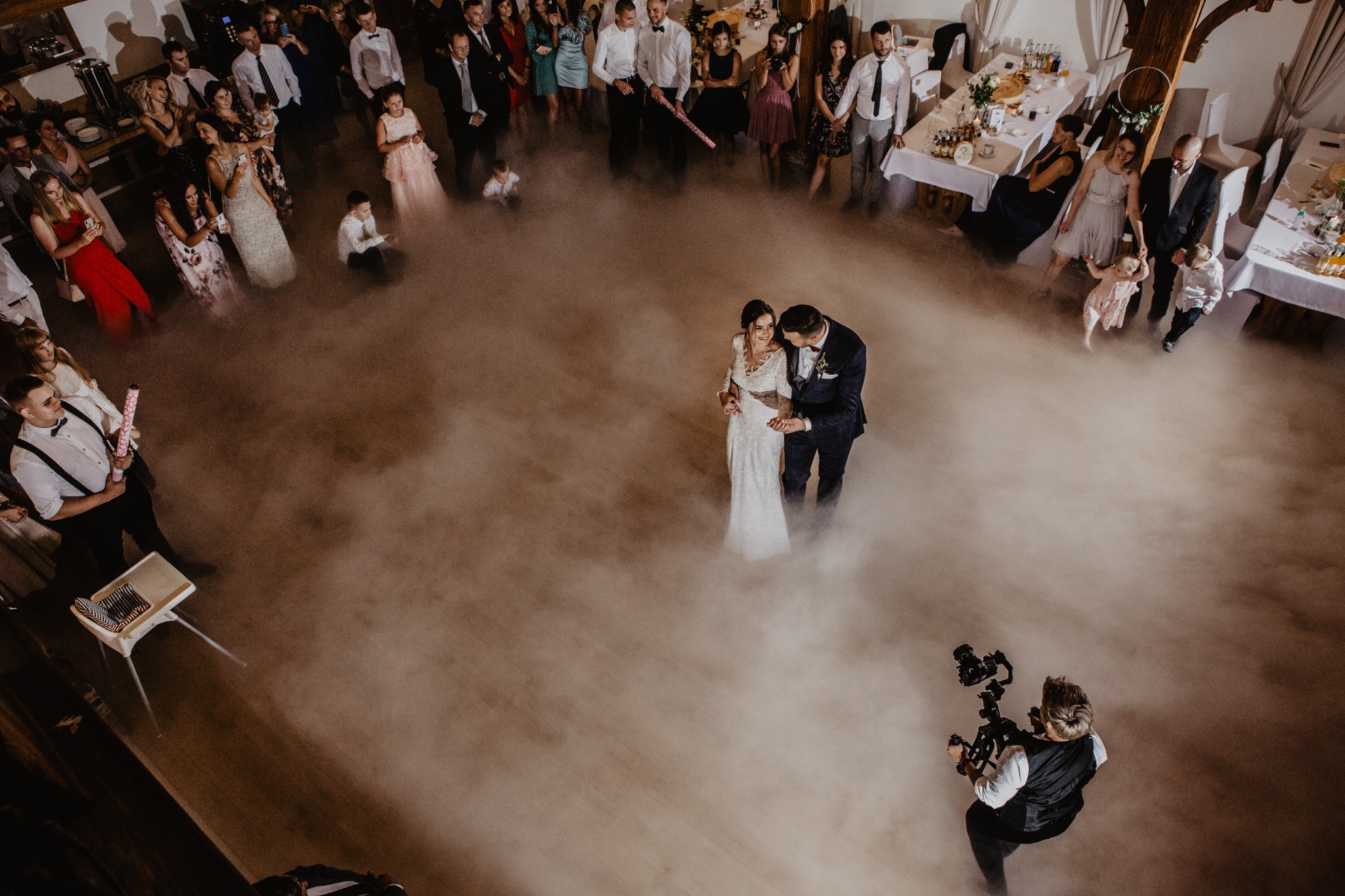 Para młoda w objęciach na parkiecie otoczonym gośćmi weselnymi i efektem mgły, filmowana z góry przez kamerzystę.