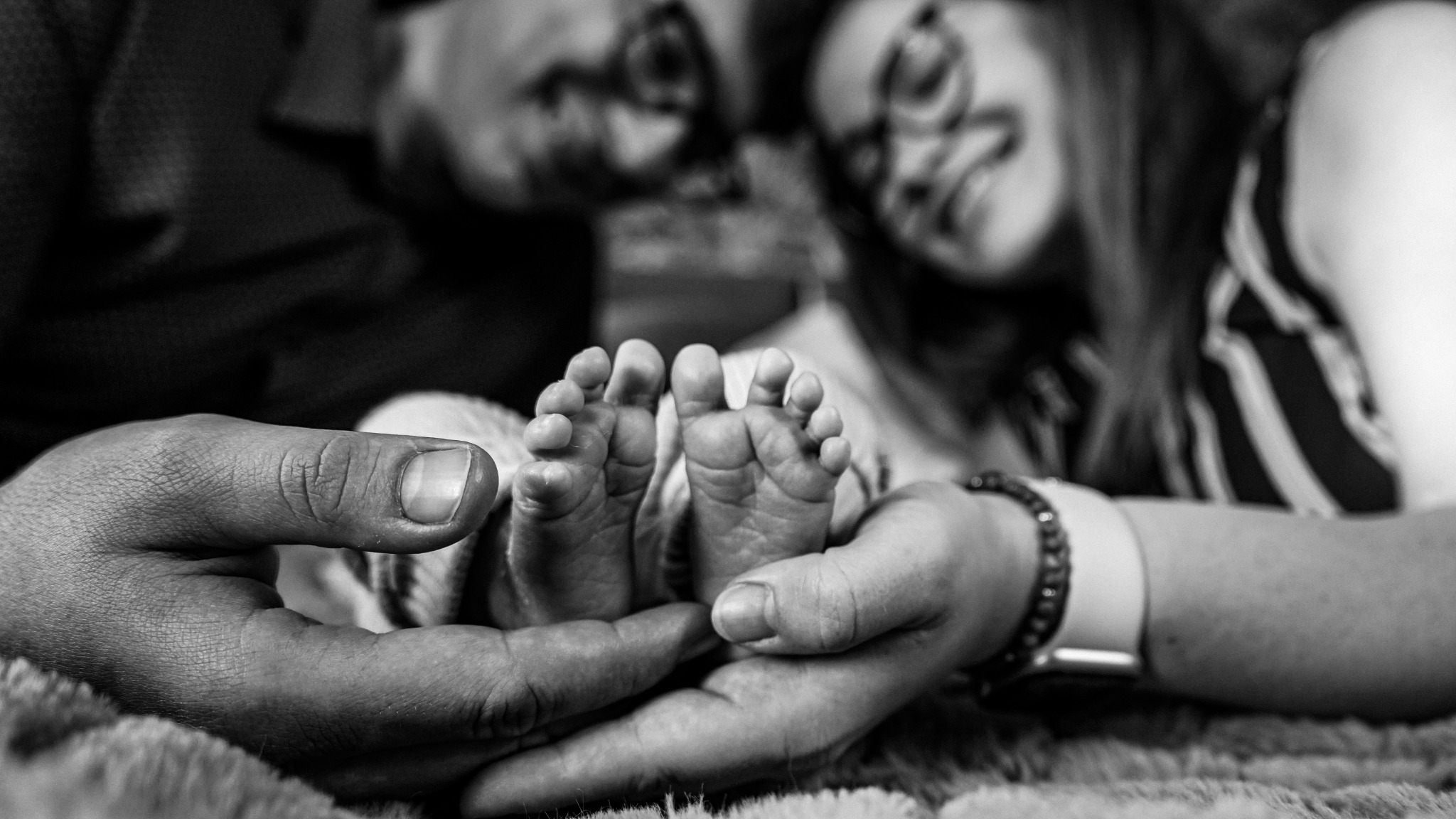 Czarno-białe zdjęcie dłoni rodziców trzymających stopy noworodka na tle rozmytych twarzy rodziców, mama ma na ręce bransoletkę i zegarek.