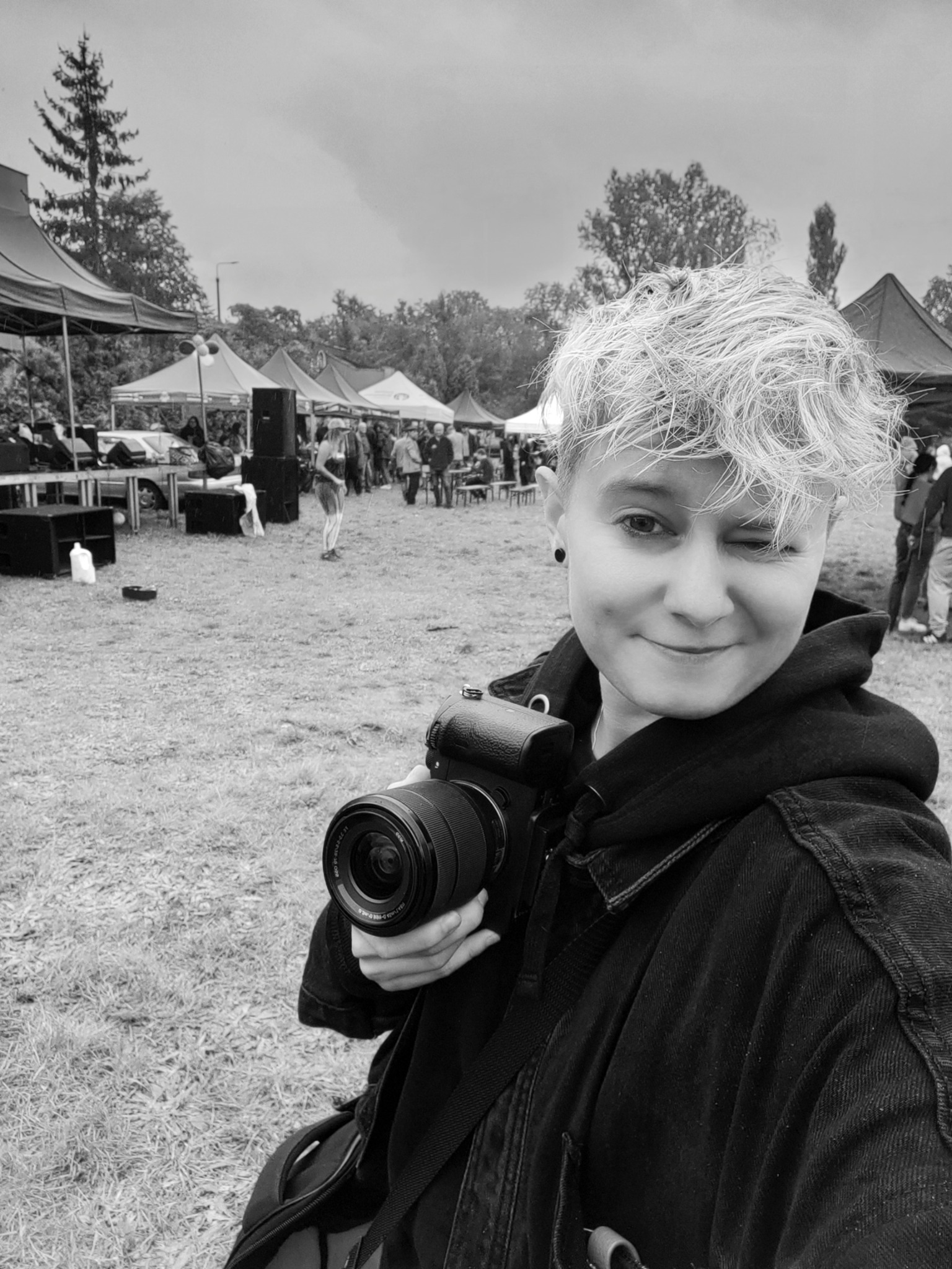 Czarno-białe zdjęcie uśmiechniętej osoby z krótkimi blond włosami mrugającej okiem, trzymającej aparat fotograficzny na tle plenerowej imprezy z namiotami i ludźmi.
