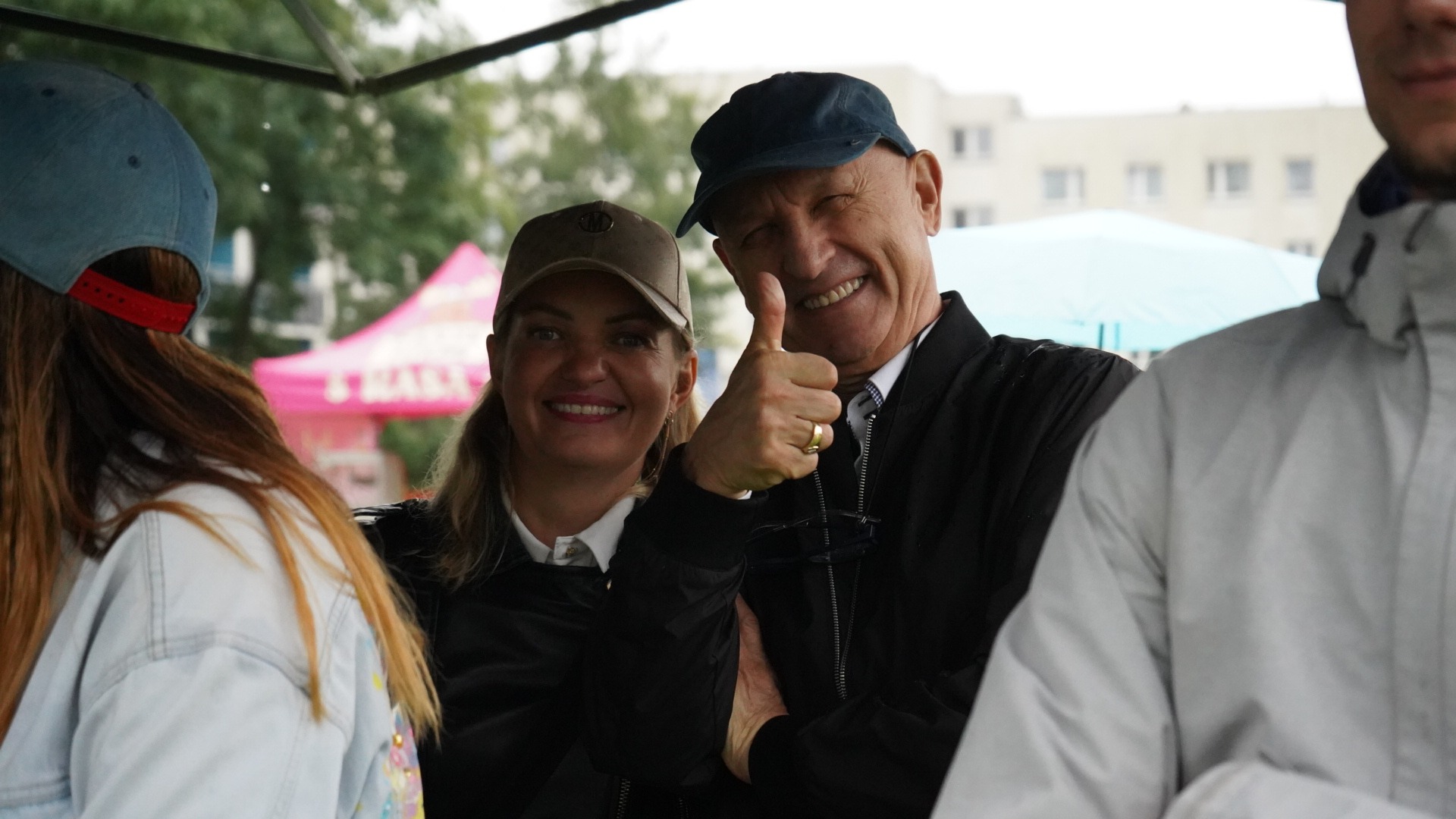 Uśmiechnięta para i mężczyzna w tle, kciuk w górę, plener, budynek w oddali.