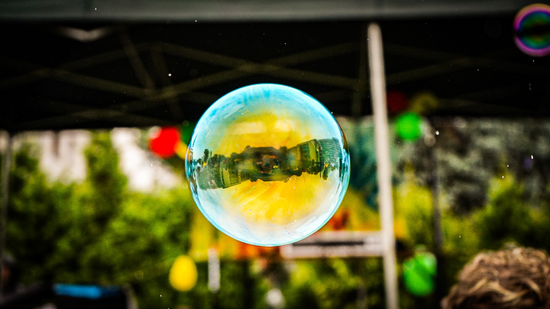 Duża bańka mydlana z odbiciem budynku na tle zieleni i kolorowych dekoracji.