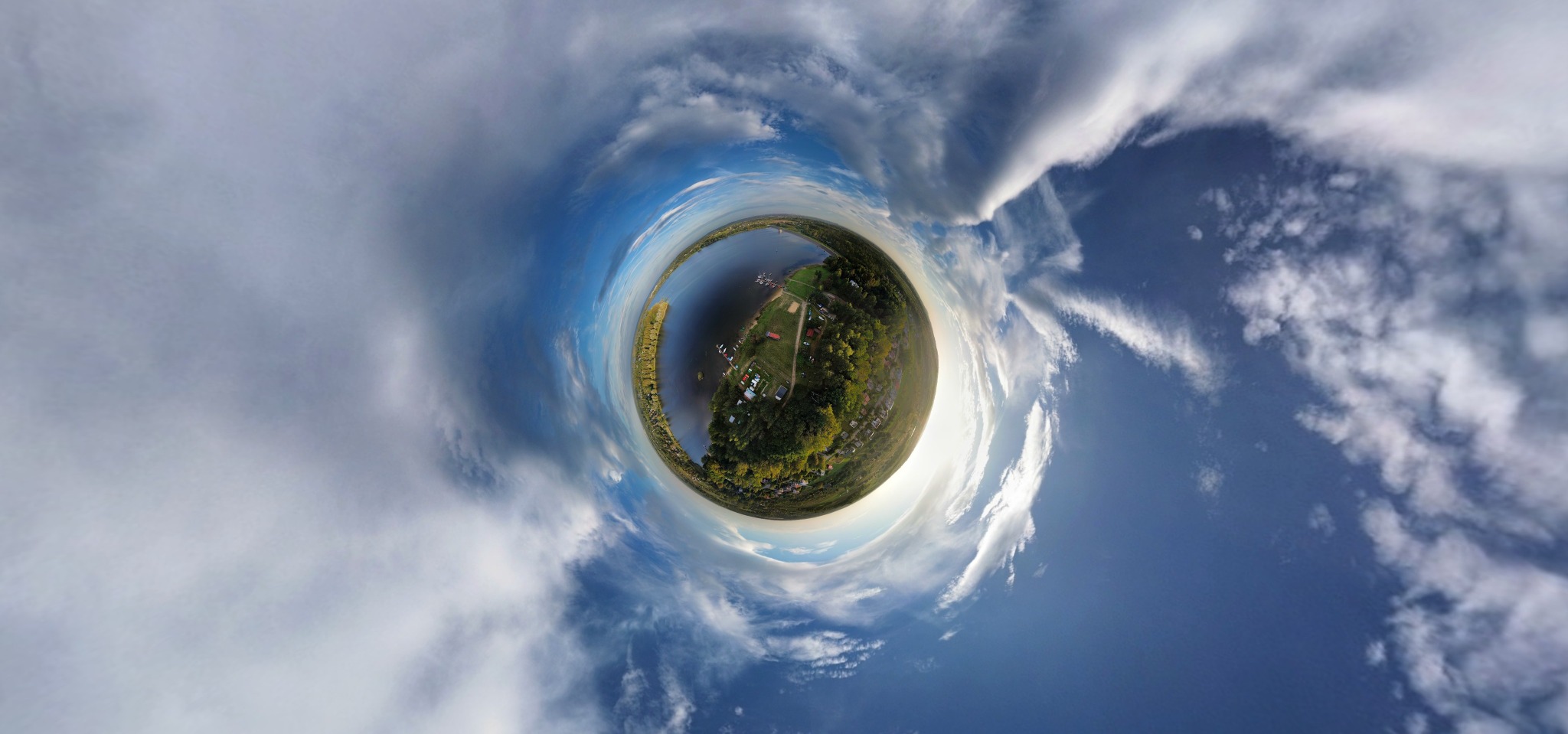 Panoramiczne ujęcie sferyczne jeziora otoczonego zielenią i zabudowaniami, z niebem i chmurami tworzącymi ramę.