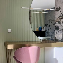 LAMAR meble - Nowoczesna toaletka z półokrągłym lustrem i różowym krzesłem na tle zielonej ściany z subtelnym wzorem. Złote detale blatu i nóg dodają elegancji.