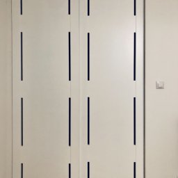 LAMAR meble - Białe, przesuwne drzwi z prostokątnymi, pionowymi wycięciami, widoczne na tle parkietu i ściany z włącznikiem światła.