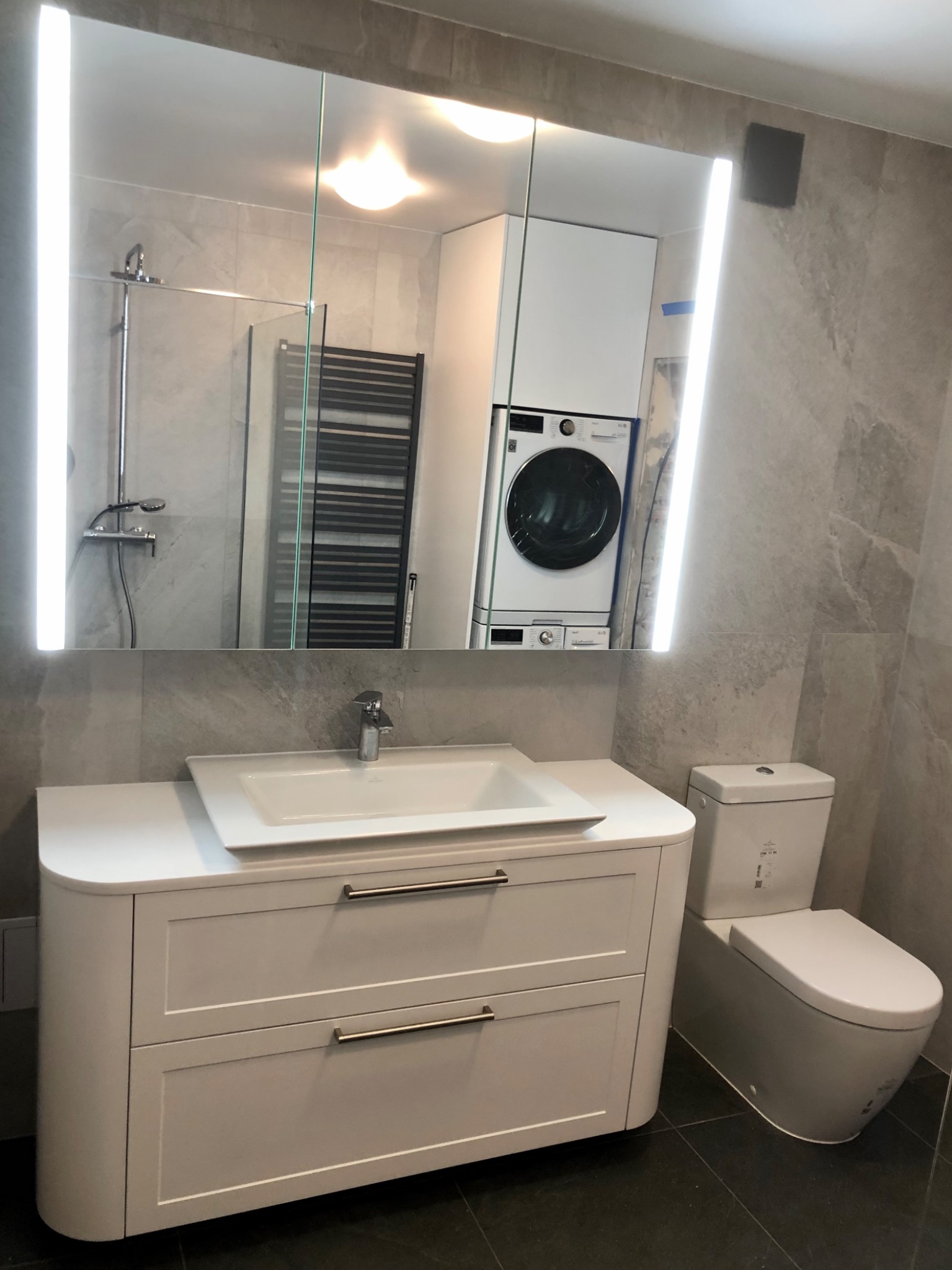 Nowoczesna łazienka z szafką pod umywalkę z szufladami, toaletą i lustrem z podświetleniem LED, w którym odbija się pralka i kabina prysznicowa.