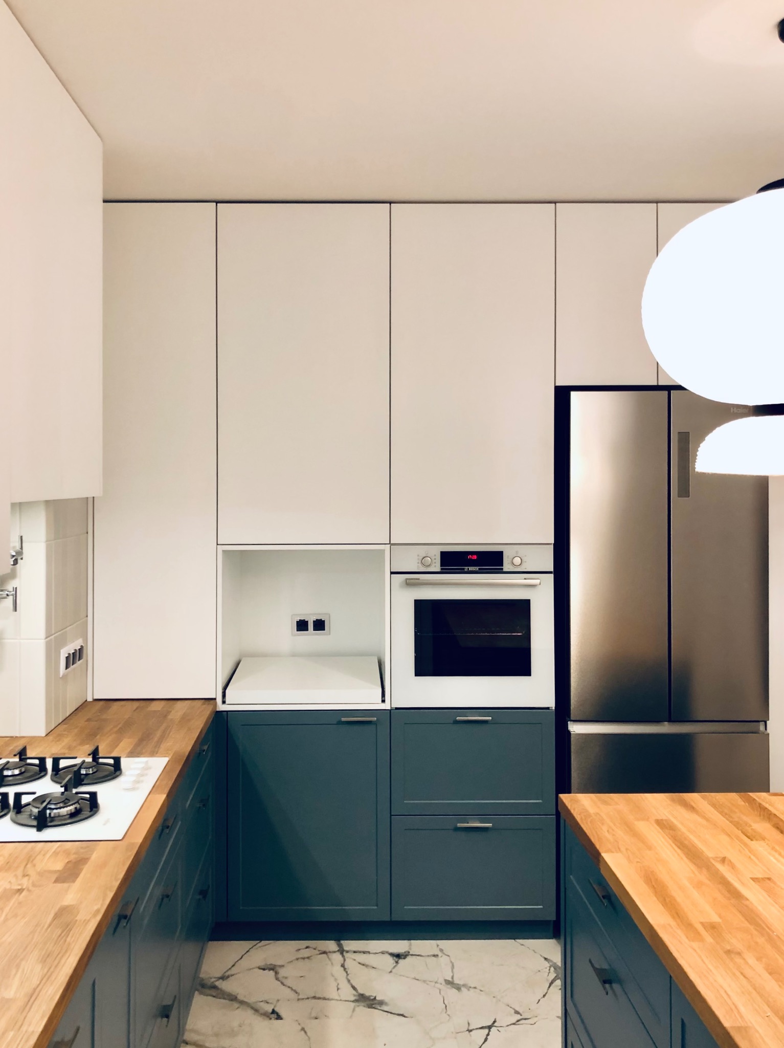 Nowoczesna kuchnia z białymi szafkami górnymi i szarymi dolnymi, drewniany blat, marmurowa podłoga. Widoczny piekarnik Bosch i lodówka side-by-side.