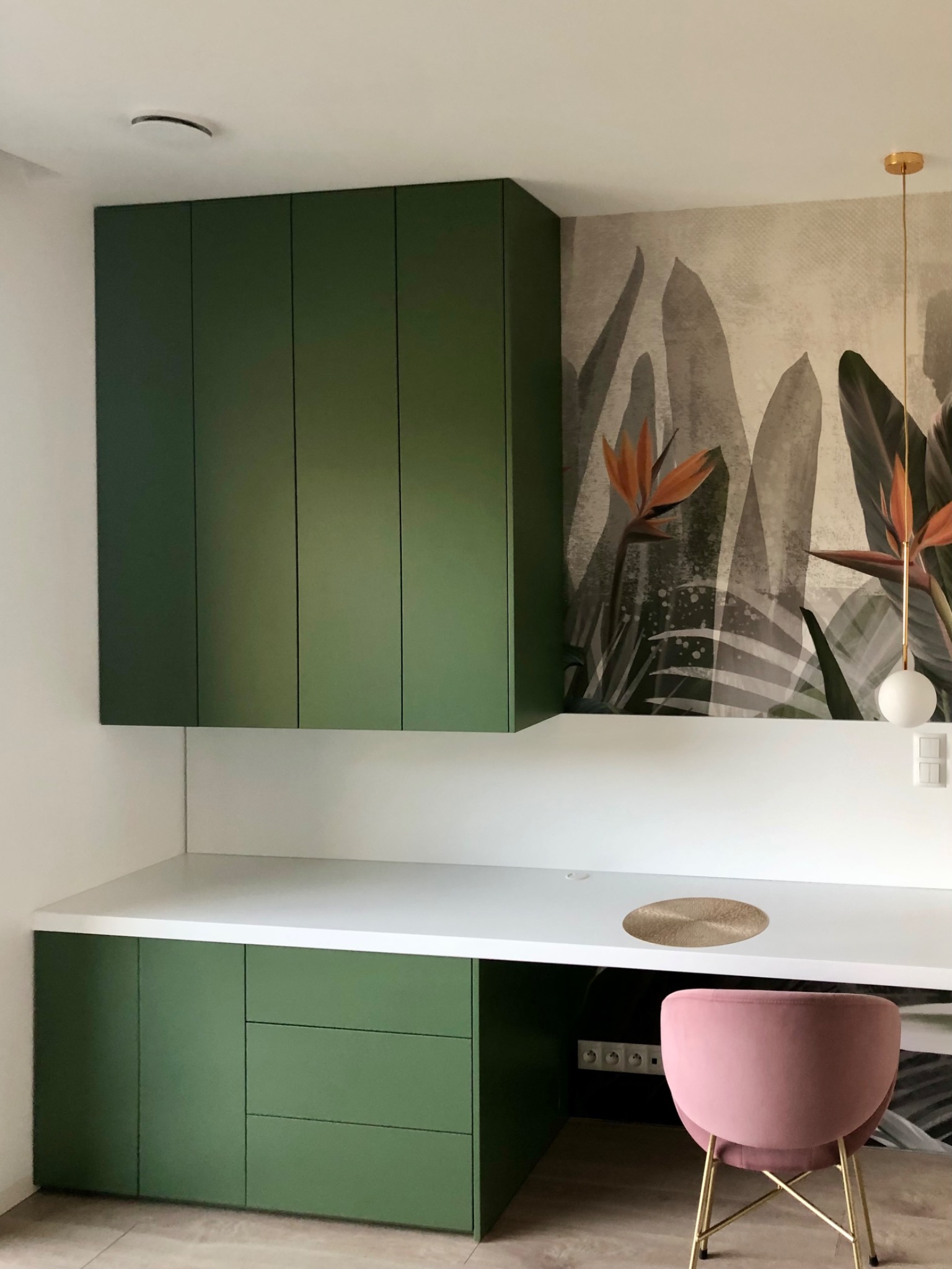 Zielone szafki kuchenne podwieszane i stojące z białym blatem, uzupełnione tapetą z motywem roślinnym i różowym krzesłem, tworzą nowoczesną przestrzeń do pracy.