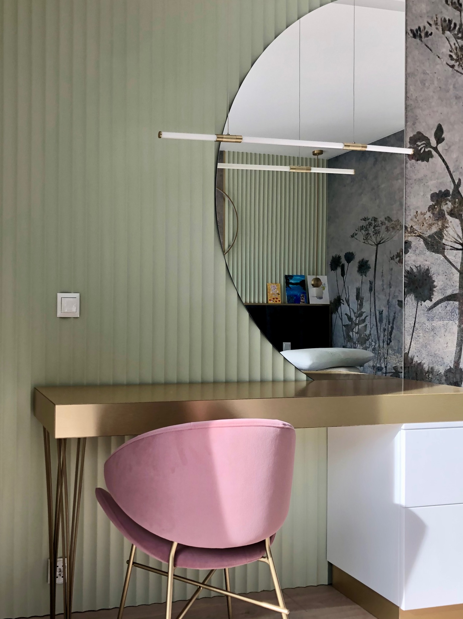 Nowoczesna toaletka z półokrągłym lustrem i różowym krzesłem na tle zielonej ściany z subtelnym wzorem. Złote detale blatu i nóg dodają elegancji.