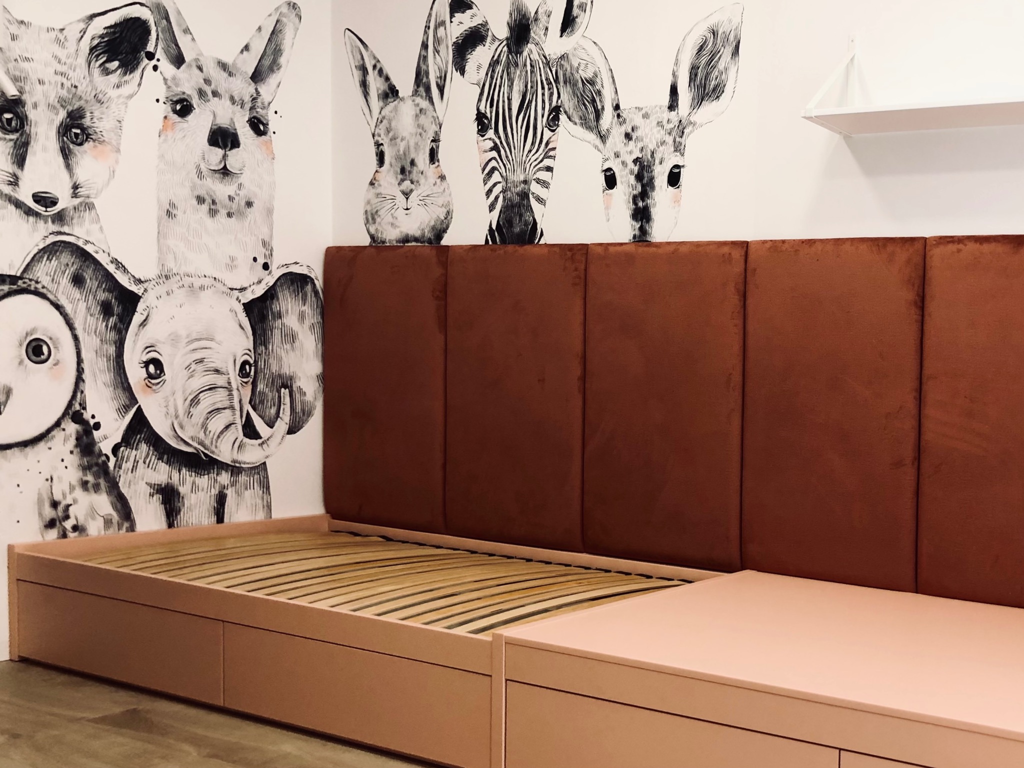 Różowe łóżko z brązowym wezgłowiem na tle ściany z rysunkami zwierząt. Nowoczesny design, widoczne detale ramy i stelaża. Ujęcie z bliska, podkreślające estetykę mebla.