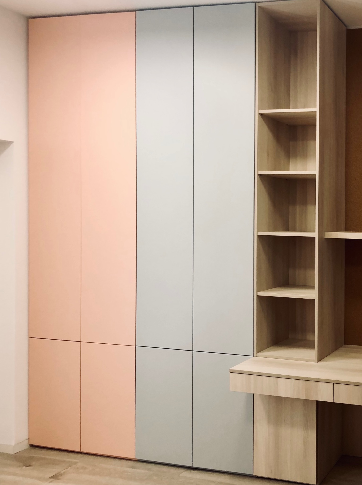 Nowoczesna szafa wnękowa z biurkiem, fronty w odcieniach różu i szarości, drewniane półki po prawej stronie. Minimalistyczny design, jasne wnętrze.