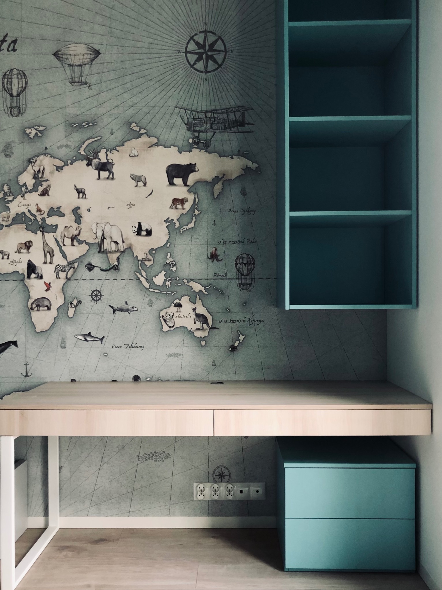 Błękitny regał wiszący nad biurkiem z jasnego drewna, na tle ściany z mapą świata w stylu vintage z motywami zwierząt i statków powietrznych.