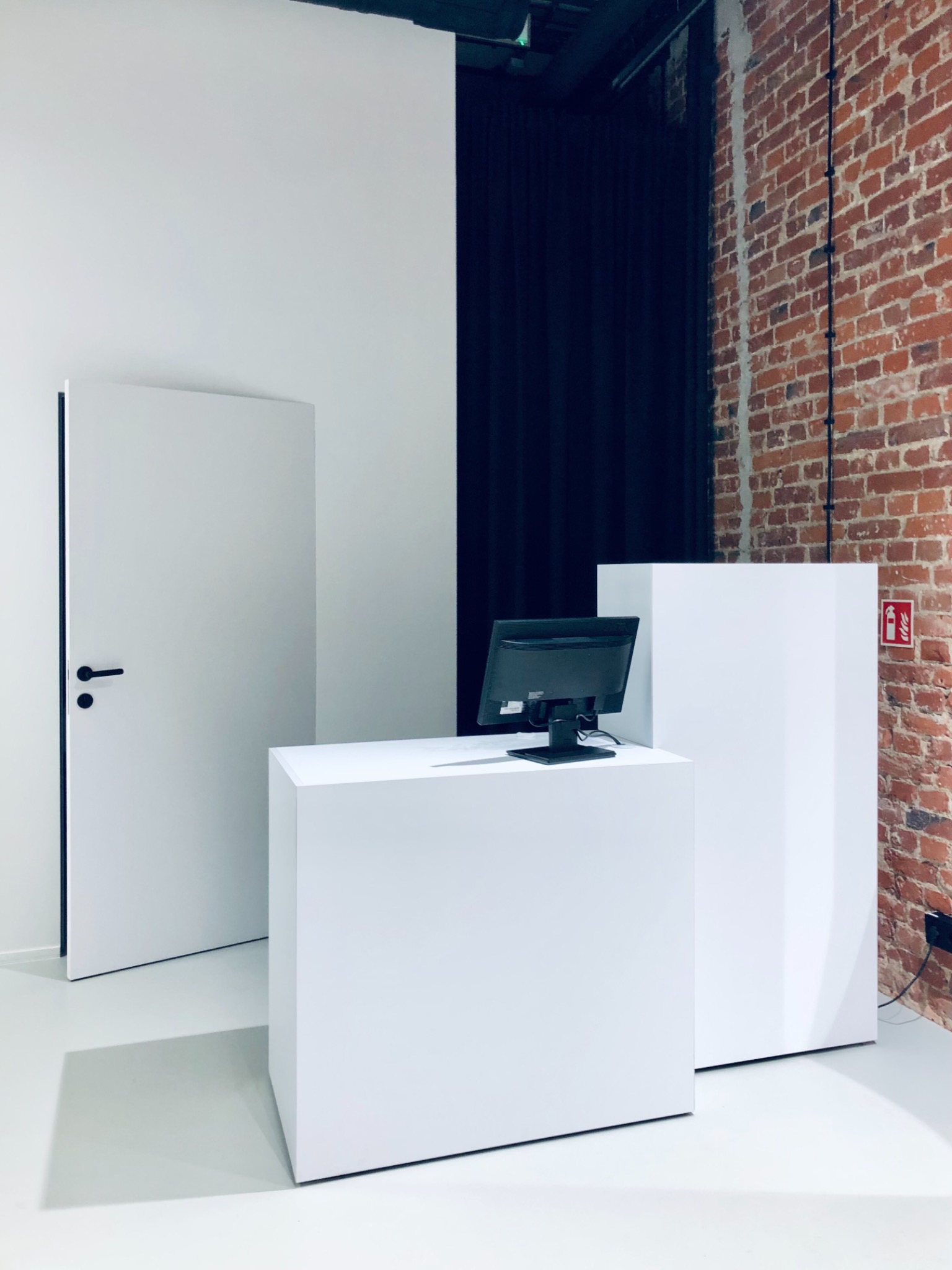 Minimalistyczne wnętrze z białą ladą recepcyjną, monitorem, drzwiami i fragmentem ściany z czerwonej cegły.