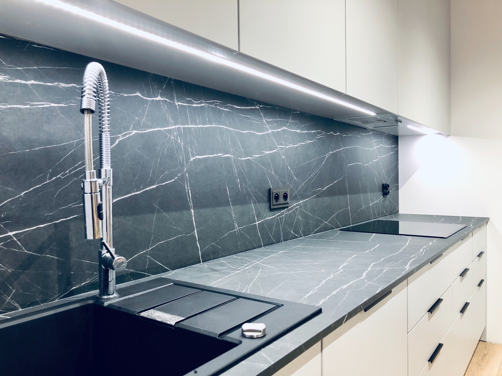Nowoczesna kuchnia z czarnym zlewem, baterią z wyciąganą wylewką i blatem imitującym marmur, podświetlona listwą LED pod szafkami.