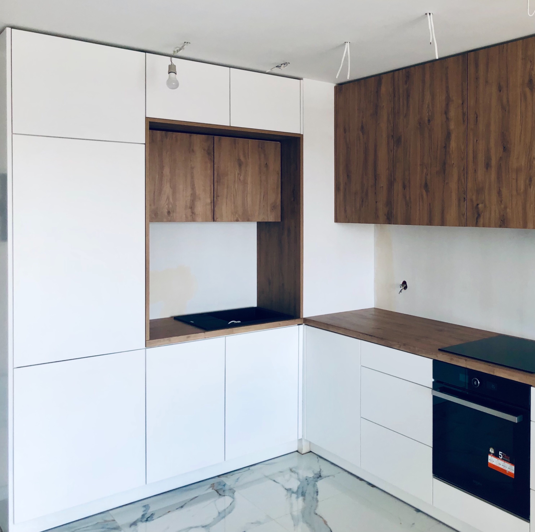 Białe i drewnopodobne szafki kuchenne z blatem, płyta indukcyjna i piekarnik, podłoga z marmurowym wzorem, widoczne przewody oświetleniowe sufitowe.