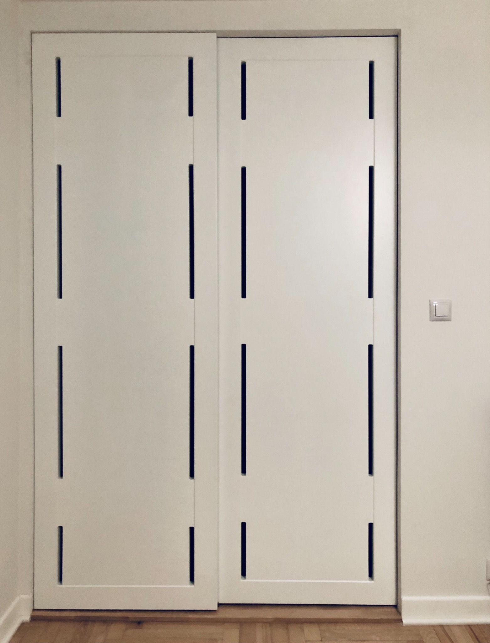 Białe, przesuwne drzwi z prostokątnymi, pionowymi wycięciami, widoczne na tle parkietu i ściany z włącznikiem światła.