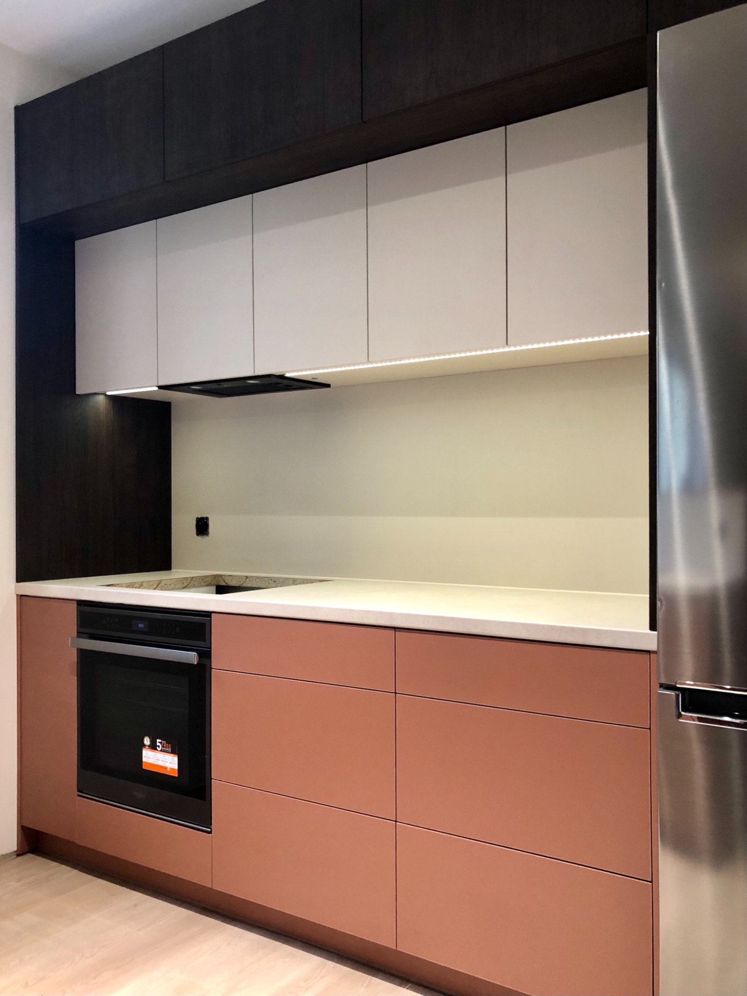 Nowoczesna kuchnia z frontami w kolorze miedzianym, jasnym blatem i białymi szafkami górnymi z oświetleniem LED, obok lodówka ze stali nierdzewnej.