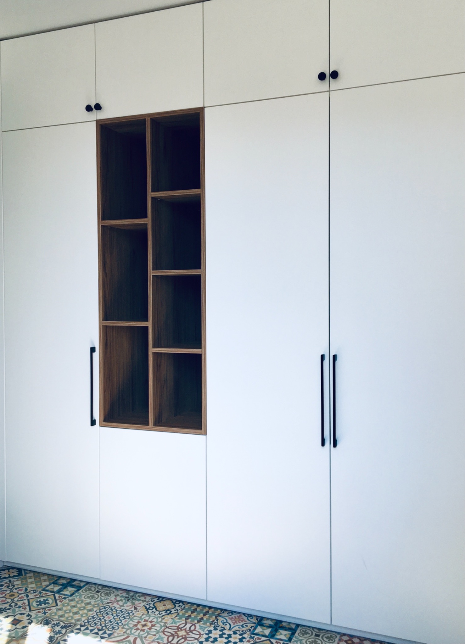 Biała szafa wnękowa z drewnianą, asymetryczną półką, czarne uchwyty, wzorzysta podłoga z płytek.