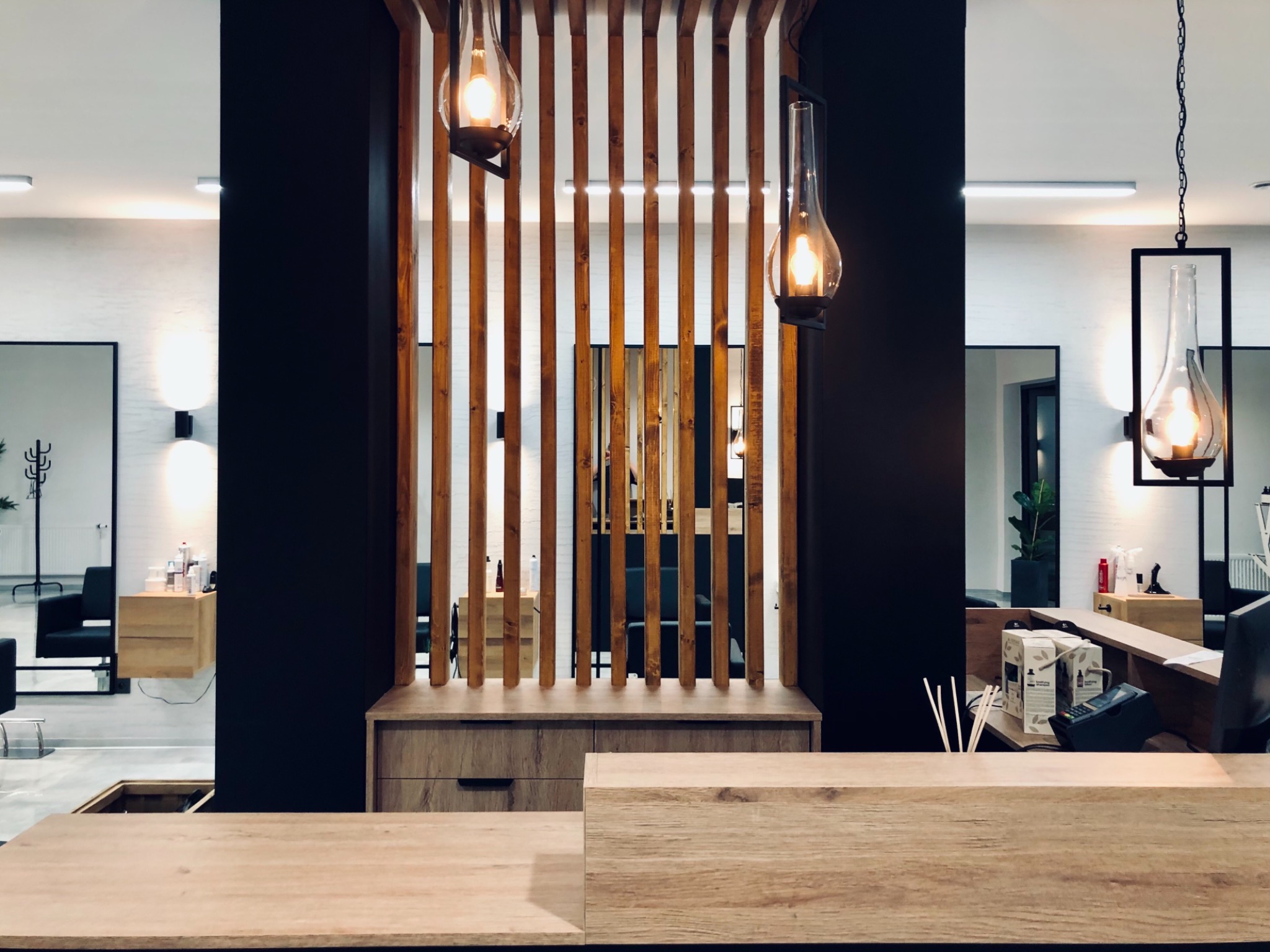 Nowoczesne wnętrze salonu fryzjerskiego z drewnianą recepcją, dekoracyjną ścianką z pionowych listew i wiszącymi lampami w stylu retro.