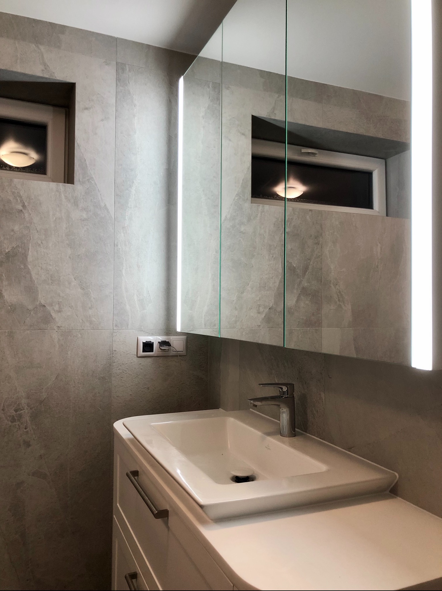 Nowoczesna łazienka z umywalką nablatową na białej szafce, szara okładzina ścienna imitująca kamień oraz lustro z podświetleniem LED, widoczne gniazdka elektryczne.