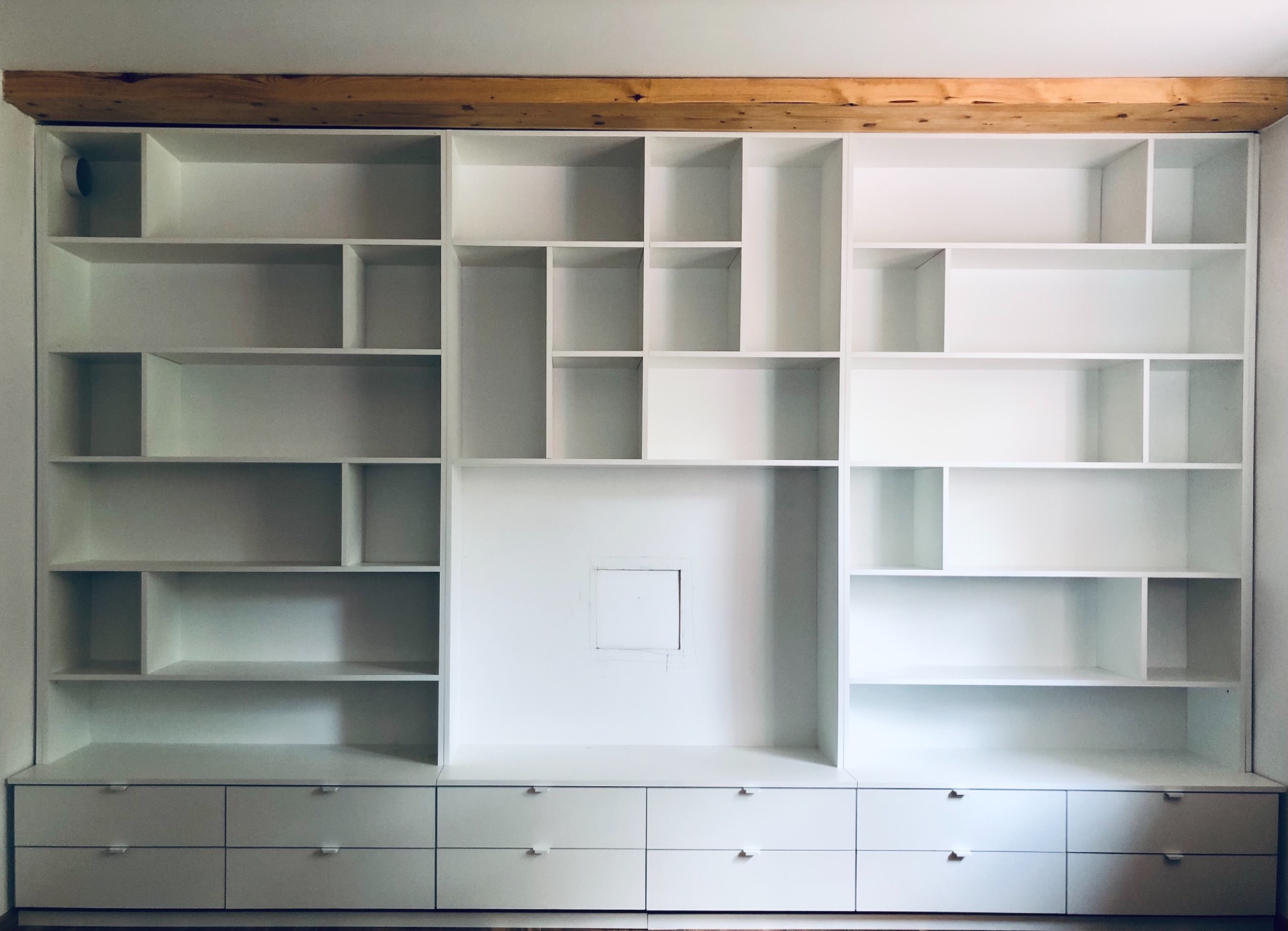 Biała, zabudowana szafa z szufladami i otwartymi półkami o nieregularnych kształtach, z drewnianą belką na górze, w minimalistycznym wnętrzu.