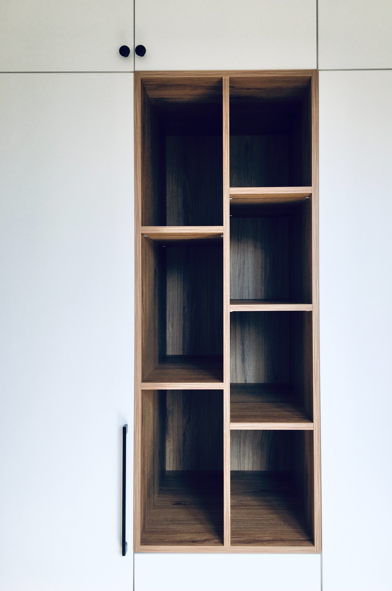 Biała zabudowa meblowa z drewnianym regałem o asymetrycznym układzie półek, czarne, minimalistyczne uchwyty.