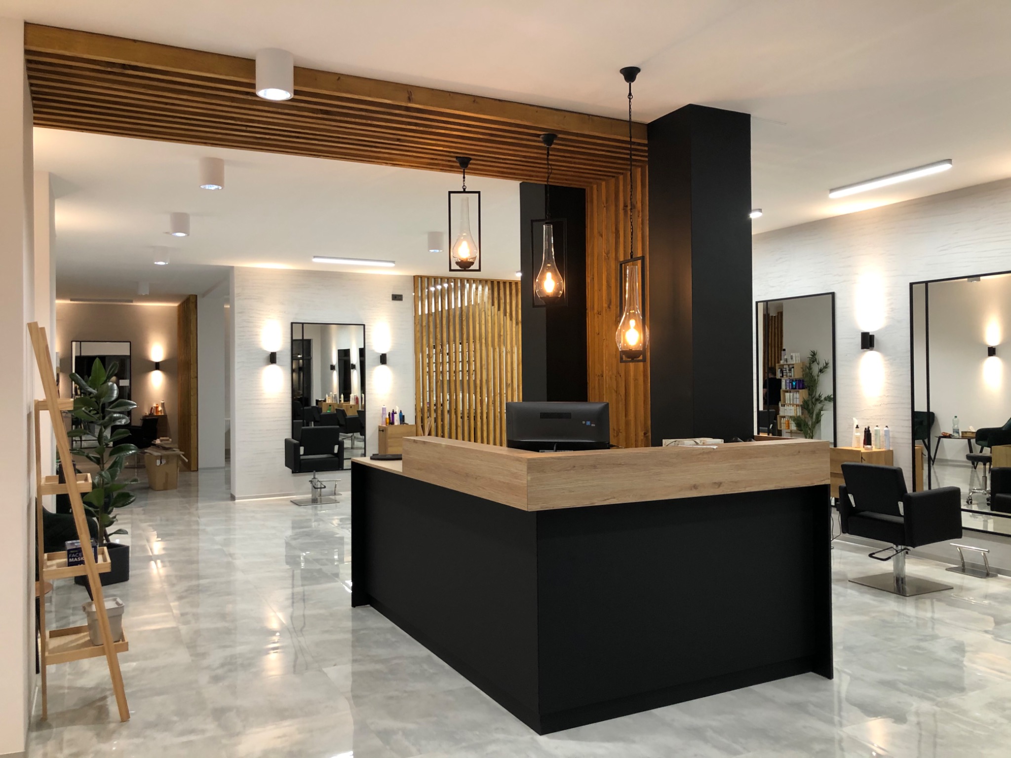 Nowoczesne wnętrze salonu fryzjerskiego z czarno-drewnianą recepcją, wiszącymi lampami w stylu loft, marmurową podłogą i drewnianymi panelami dekoracyjnymi.