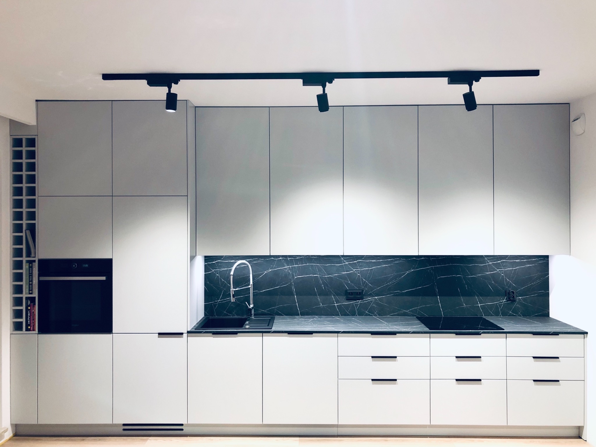 Nowoczesna, minimalistyczna kuchnia z białymi, matowymi frontami, czarnym blatem z marmurowym wzorem i czarnymi, punktowymi lampami sufitowymi.