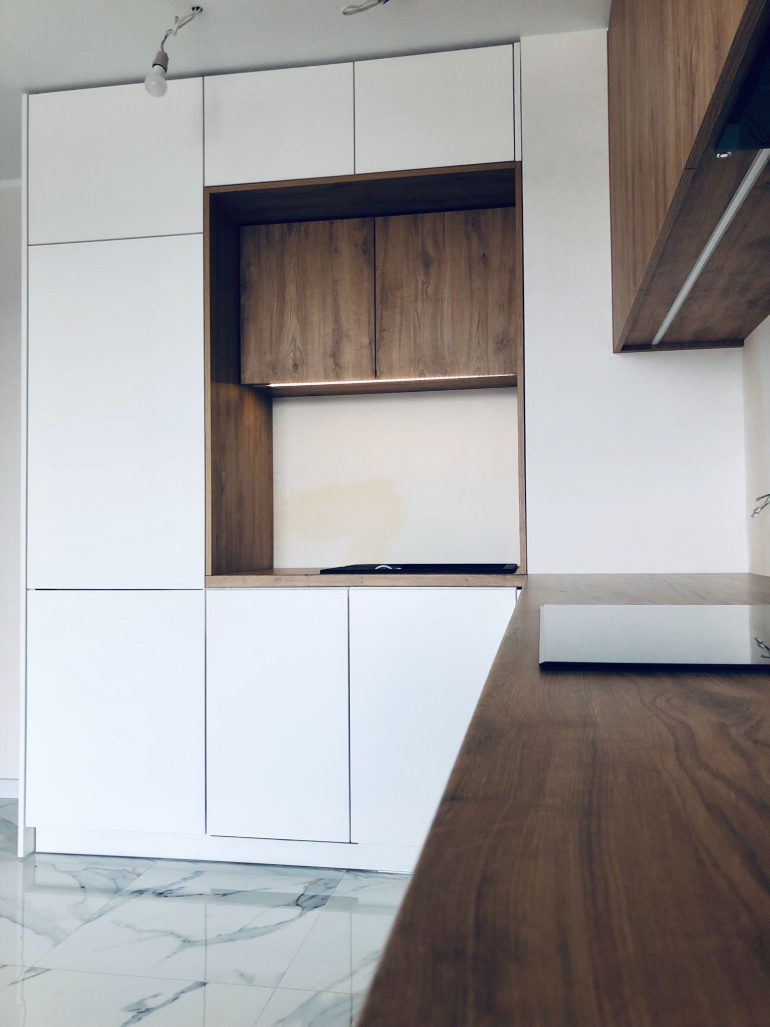 Nowoczesna kuchnia w Łodzi z białymi frontami i drewnianymi akcentami, oświetlenie LED w półce, marmurowe płytki podłogowe.