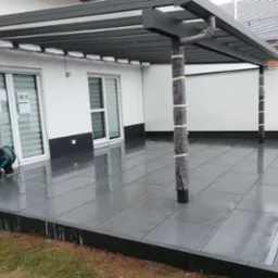 Nowoczesny taras z ciemnych płyt, zadaszony pergolą z szarymi belkami, zabezpieczone filcem słupy, widoczny zwinięty wąż ogrodowy przy ścianie budynku.