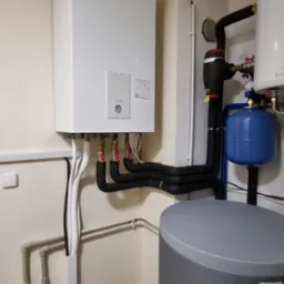 Biała jednostka wewnętrzna Panasonic z podłączonymi rurami, zbiornik buforowy w kolorze szarym, instalacja w pomieszczeniu technicznym z beżowymi ścianami i drewnopodobną podłogą.