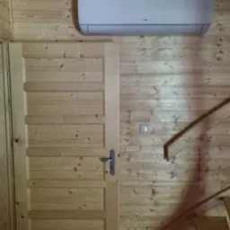 Wnętrze domku z drewnianą boazerią: biała jednostka klimatyzacyjna AUX zamontowana nad drewnianymi drzwiami, obok schodów i włącznika światła.