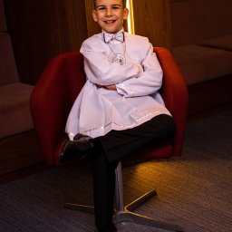 Szymon Roman - Uśmiechnięty chłopiec w stroju komunijnym, siedzący na czerwonym krześle. Elegancka koszula, mucha i medalik. Ciepłe oświetlenie i nowoczesne wnętrze w tle.