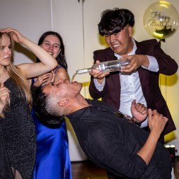 Szymon Roman - Mężczyzna pije alkohol wlewany mu do ust przez kolegę, podczas gdy dwie kobiety się śmieją. W tle balon z napisem 'Happy Birthday'.