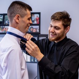 Szymon Roman - Uśmiechnięty mężczyzna w czarnej koszuli poprawia krawat drugiemu mężczyźnie w białej koszuli. W tle półka z ramkami ze zdjęciami.