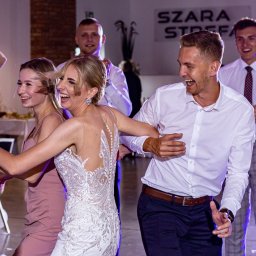 Szymon Roman - Rozbawiona para młoda w tańcu z gośćmi weselnymi. Panna młoda w koronkowej sukni, pan młody w białej koszuli. W tle uśmiechnięci goście i dekoracje sali.