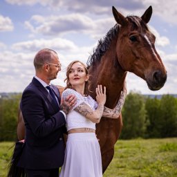 Szymon Roman - Para młoda w objęciach z koniem na tle łąki i błękitnego nieba z chmurami. Pan młody w garniturze, panna młoda w białym stroju. Sesja plenerowa z zwierzęciem.