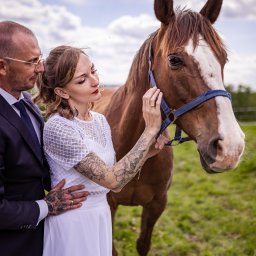 Szymon Roman - Para młoda w plenerze z koniem. Panna młoda w białej sukience z tatuażami, pan młody w garniturze. Romantyczna sesja w naturalnym otoczeniu, zieleń i błękitne niebo w tle.