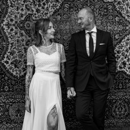 Szymon Roman - Czarno-białe zdjęcie pary młodej, opierającej się o wzorzystą ścianę. Ona w sukni z rozcięciem i tatuażem na udzie, on w garniturze, trzymają się za ręce i patrzą na siebie.
