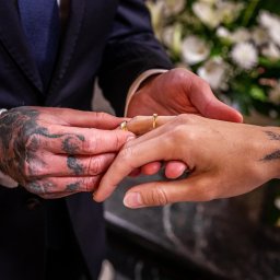 Szymon Roman - Tatuowane dłonie pary młodej podczas ceremonii ślubnej: on zakłada obrączkę jej na palec, w tle bukiet białych kwiatów.