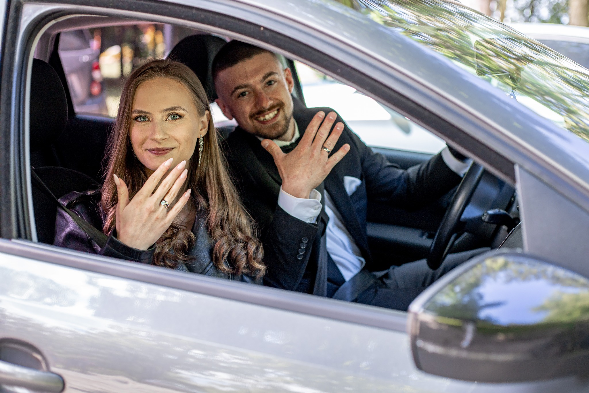 Para młoda w samochodzie prezentuje obrączki ślubne. Ona w skórzanej kurtce, on w garniturze. Uśmiechnięci, radośni. Widok z zewnątrz samochodu.
