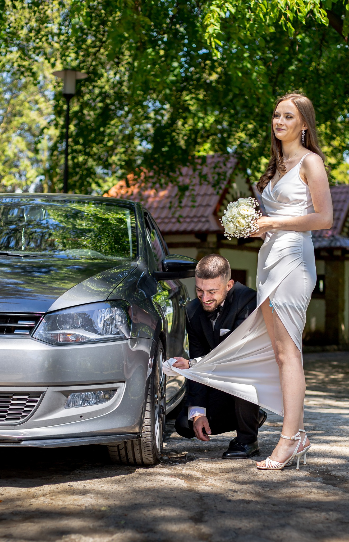 Uśmiechnięty pan młody w czarnym smokingu czyści suknię panny młodej przy srebrnym samochodzie, ona trzyma bukiet białych róż i patrzy w dal. Słoneczny dzień.