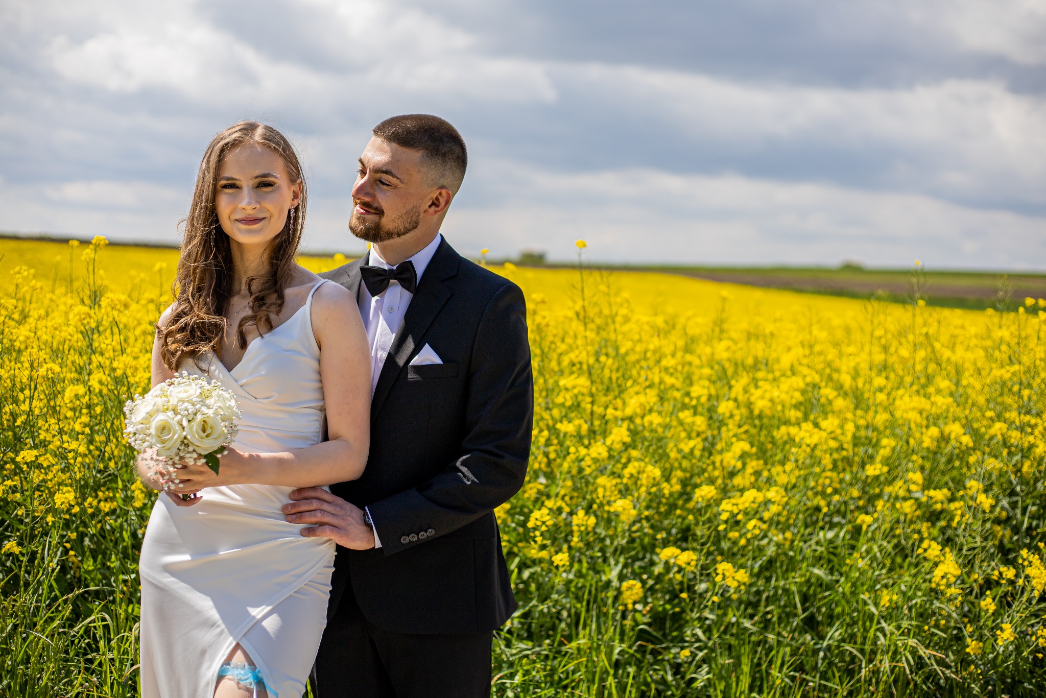 Para młoda w plenerze kwitnącego rzepaku. Panna młoda trzyma bukiet białych róż, widoczna podwiązka. Pan młody w czarnym garniturze, patrzy na pannę młodą.
