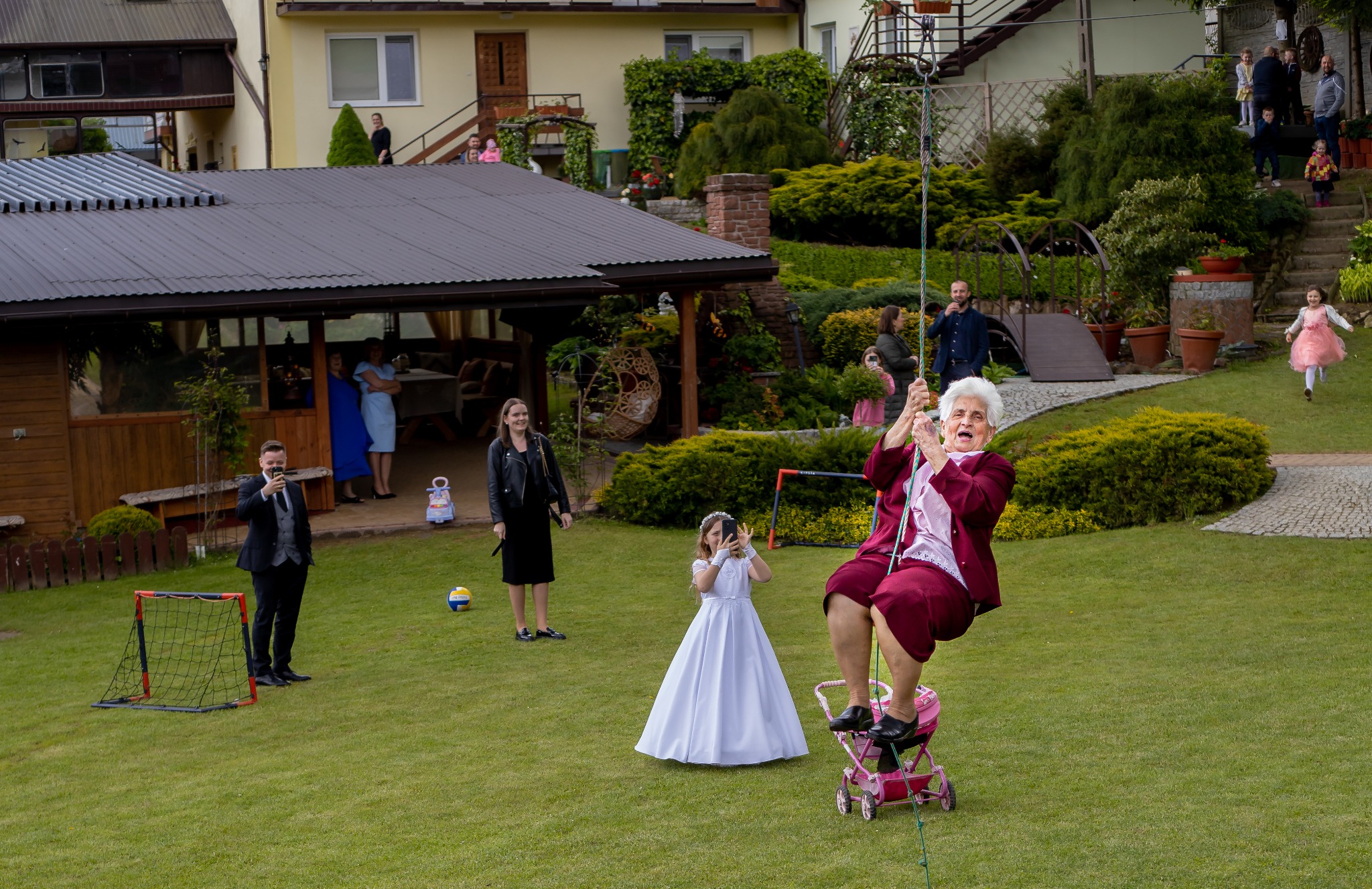Radosna seniorka w bordowym stroju zjeżdża na huśtawce, otoczona przez fotografa i dziewczynkę w białej sukience na tle bujnego ogrodu podczas uroczystości rodzinnej.