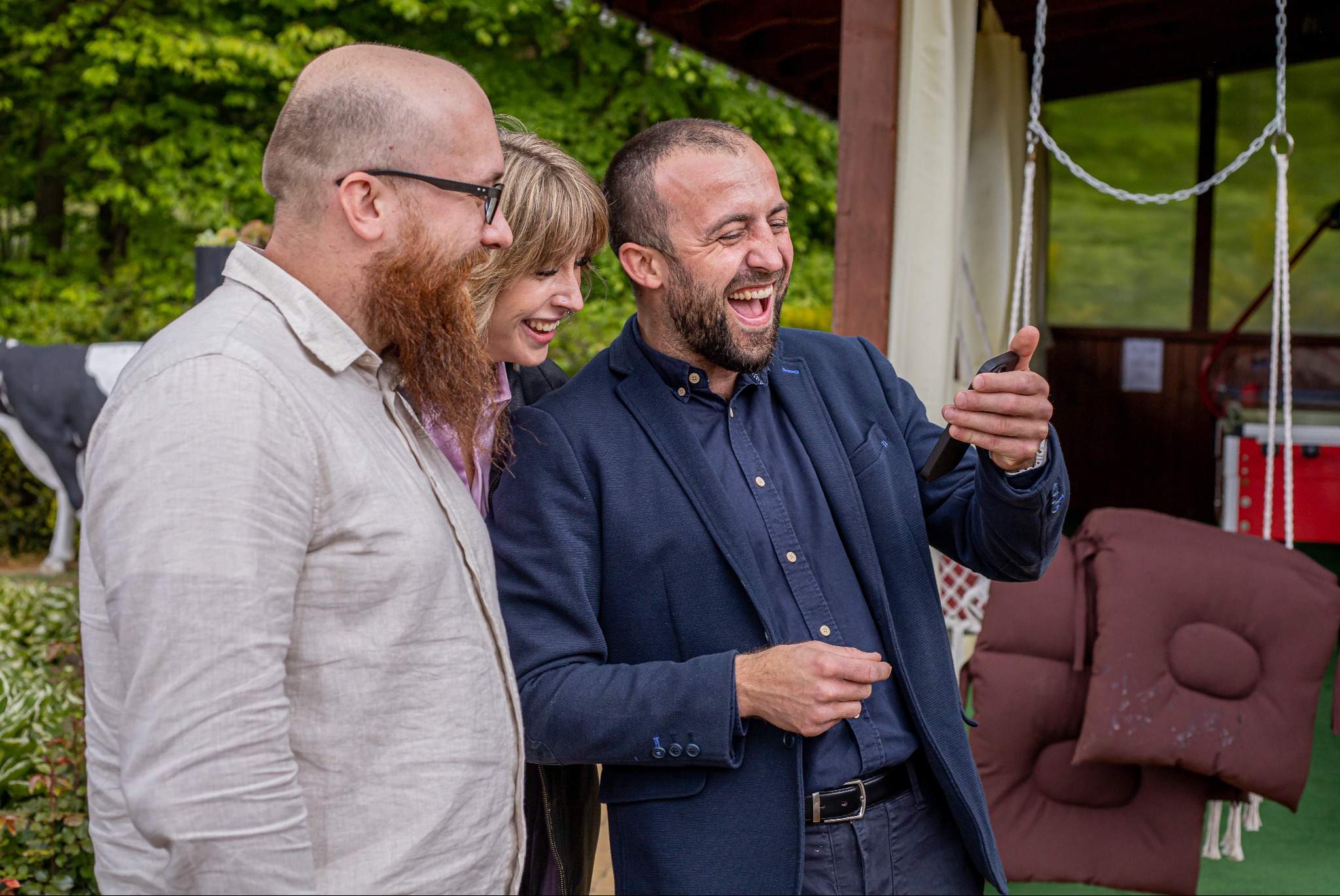 Radosne chwile: Trzy osoby śmieją się, patrząc na telefon. Mężczyzna w niebieskiej marynarce trzyma urządzenie. W tle zielona roślinność i huśtawka.