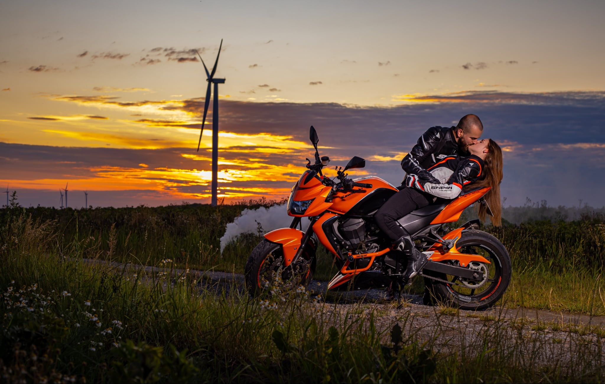 Para w skórzanych strojach motocyklowych całuje się na pomarańczowym motocyklu na tle zachodzącego słońca i turbin wiatrowych. Romantyczna sesja plenerowa.