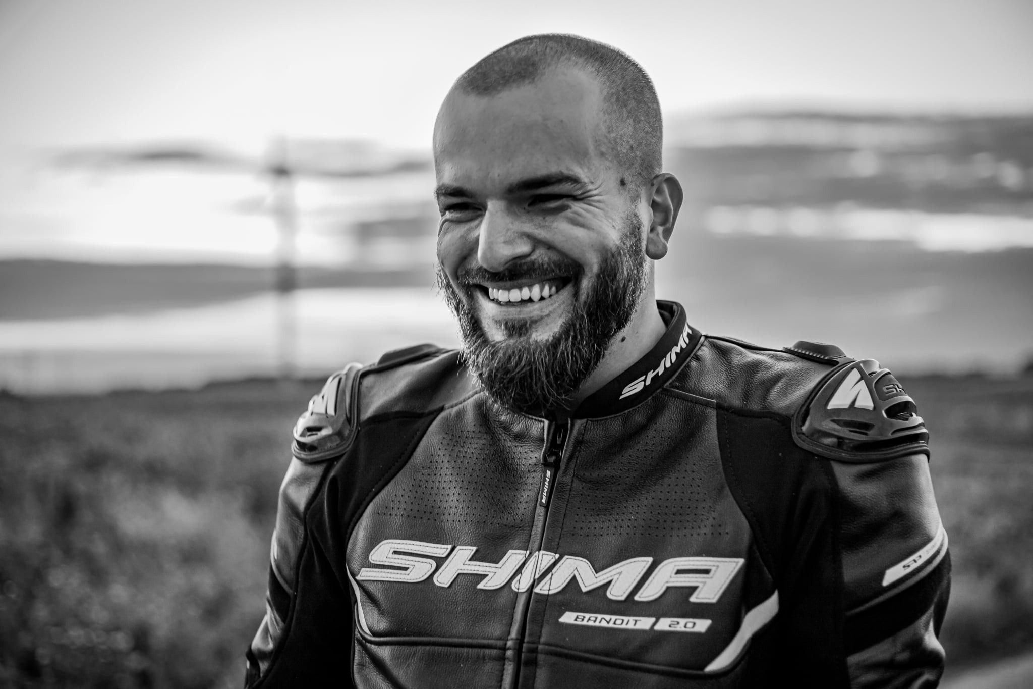 Czarno-białe zdjęcie uśmiechniętego mężczyzny z brodą w skórzanej kurtce motocyklowej marki Shima Bandit 2.0 na tle krajobrazu. Portret w plenerze.