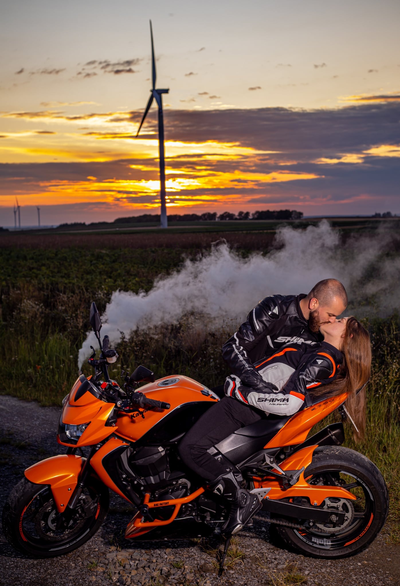 Para całująca się na pomarańczowym motocyklu Kawasaki Z750, w tle zachód słońca i turbiny wiatrowe. Sesja plenerowa z użyciem dymu.